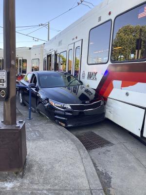 Black Kia sedan wedged between a MAX light rail train and a curb, heavy damage visible. Photo courtesy Portland Police Bureau