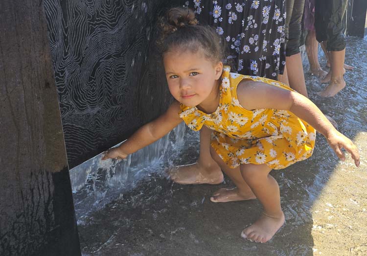 Avayah Kaahanui gets her hand wet in the shadow of the water wall at the Vancouver Waterfront Park on Saturday. Avayah was with her family, from Ridgefield, trying to cool off a bit during the heatwave. Photo by Paul Valencia